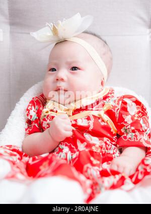 Closeup cute newborn baby in red bodysuit lying down alone on bed. Adorable infant rests on white bedsheets, staring at camera looking peaceful. Infan Stock Photo