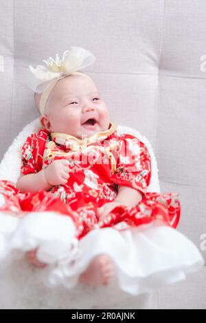 Closeup cute newborn baby in red bodysuit lying down alone on bed. Adorable infant rests on white bedsheets, staring at camera looking peaceful. Infan Stock Photo