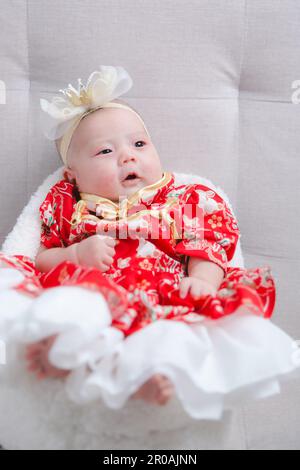 Closeup cute newborn baby in red bodysuit lying down alone on bed. Adorable infant rests on white bedsheets, staring at camera looking peaceful. Infan Stock Photo