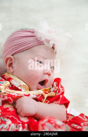 Closeup cute newborn baby in red bodysuit lying down alone on bed. Adorable infant rests on white bedsheets, staring at camera looking peaceful. Infan Stock Photo