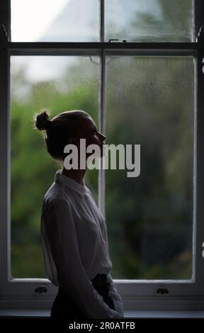 Reflection by the window. A shadowed profile view of a beautiful young woman looking away. Stock Photo