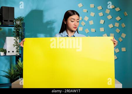 Optimistic asian woman holding yellow cardboard sign while posing in modern office on blue background. Confident female employee showing advertising poster with copy space. Stock Photo