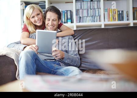 Using their digital tablet to talk to friends. an attractive young woman embracing her boyfriend while he uses a digital tablet. Stock Photo