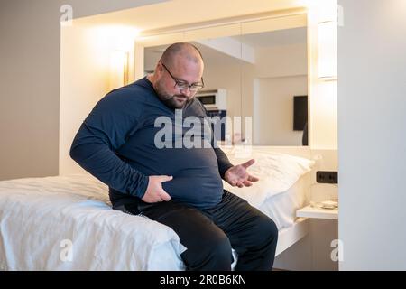 Huge man suffering from extra weight touching stomach sitting on bed in bedroom.  Stock Photo