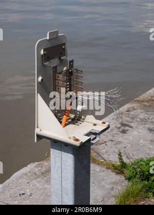 A broken pole of a barrier where the electricity wires are exposed on the side of the water Stock Photo