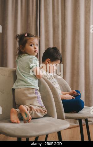 Baby girl sitting next to her brother who is playing games on the mobile phone, being obsessed with devices. Children playing video games on gadget Stock Photo