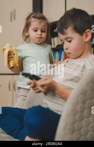 Baby girl eating a banana while watching her brother playing games on the phone. Internet technology concept. Communication concept. Education Stock Photo