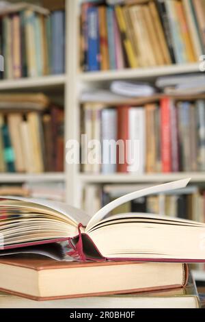 Open book or textbook on a pile of closed books in front of a bookcase in a library. Education learning concept. Vertical image with selective focus. Stock Photo