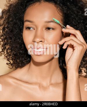 Eyebrow brushing process. Pretty curly woman combing her eyebrows with brush tool, isolated on pastel background Stock Photo