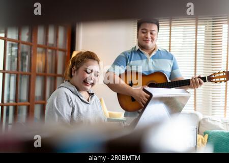 Young Asian chubby couple singing and playing acoustic guitar and piano together. Man and woman enjoying musical instrument. People in a band practicing in the house Stock Photo