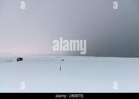 Panoramic view over a barren almost flat white snow landscape in Iceland with one car on invisible road and sky color ranging from light to dark grey Stock Photo
