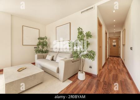 Living room in solid cream tones with a sofa flanked by artificial plants next to a long corridor Stock Photo