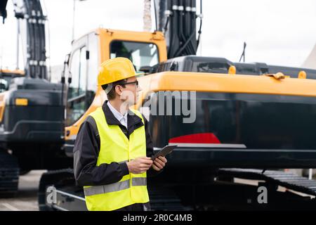 Engineer in a helmet with a digital tablet stands next to construction excavators. High quality photo Stock Photo