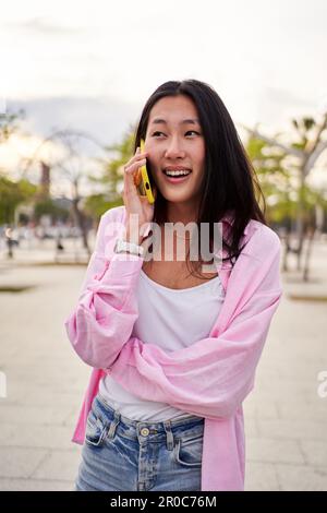 Pretty Chinese woman using smartphone being on a call while standing outdoors during summer. Stock Photo
