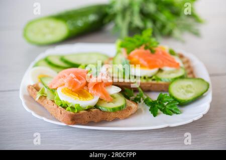 fried toast with lettuce, egg, cucumbers and red fish in a plate, on a wooden table. Stock Photo