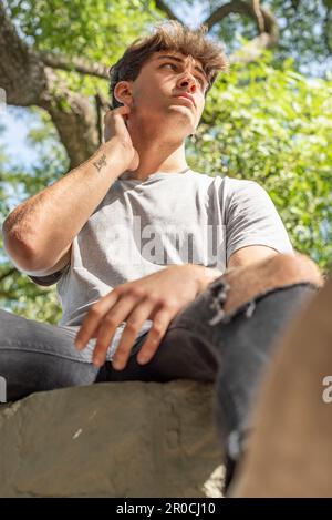 Low angle view of a young man, serious, sitting on a tree while looking away Stock Photo