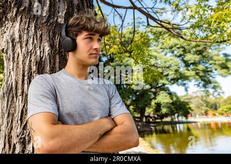 A young man listening to music, leaning on a tree with his arms folded Stock Photo
