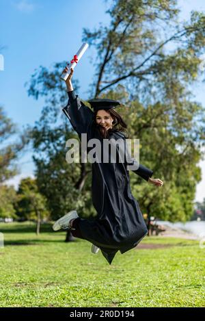 Happy graduated girl in a black gown jumping while holding her diploma Stock Photo