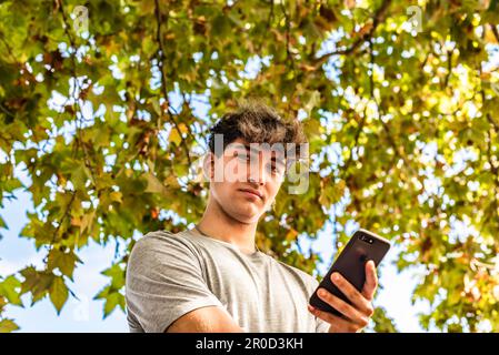 Low angle of a young man holding his smartphone, serious, looking at the camera in a public park Stock Photo