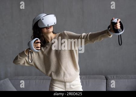A woman in virtual reality glasses with joysticks in her hands, sitting on a couch and traveling through a meta universe. VR helmet for entertainment, Stock Photo