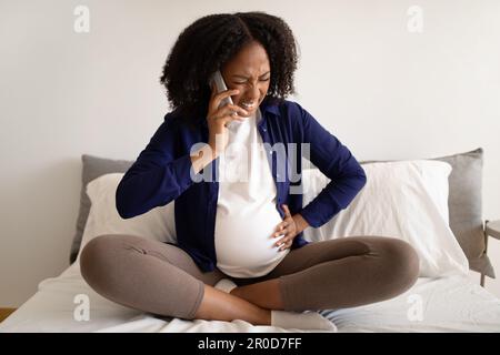 Sad millennial african american lady presses hand to belly making phone call to clinic, suffering from contractions Stock Photo