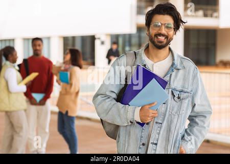 Cheerful handsome middle eastern guy in casuals and glasses with book, ready to go to lesson, at campus Stock Photo