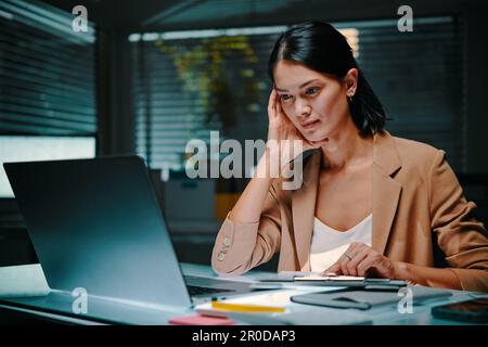 Tired businesswoman working late, reading reports and documents in laptop Stock Photo