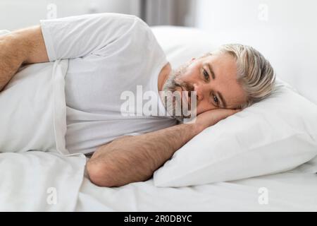 Upset grey-haired man lying in bed in the morning Stock Photo
