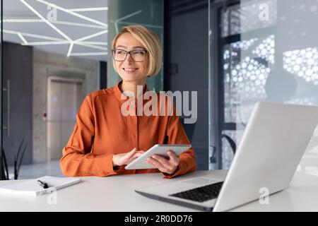 Young successful businesswoman at workplace is satisfied with results of achievement at work smiling and using tablet computer, blonde programmer is testing new software in application Stock Photo