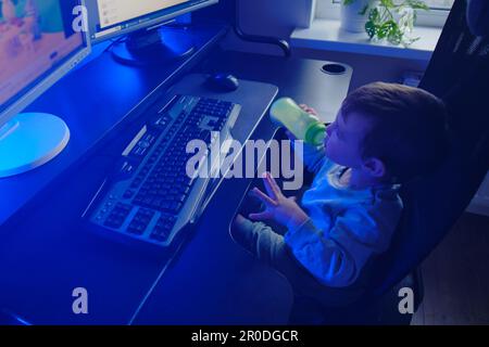 Happy baby is sitting at the computer keyboard with a bottle of milk in his hands. A child with a desktop PC in a home living room drinks milk formula Stock Photo
