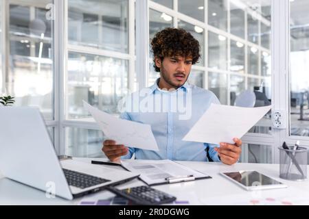 Pensive serious businessman reading financial report, hispanic businessman holding document in hands looking disappointed, working inside modern office with laptop behind paper work. Stock Photo