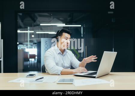 Portrait of a young Asian man in a headset. Sits in the office at the laptop and consults, talks through a video call from the laptop. Stock Photo