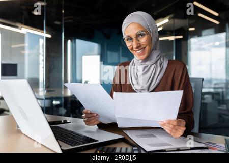 Portrait of young smiling Muslim designer woman in hijab smiling at camera, working in office at laptop and with documents, graphs, drawings, projects. Stock Photo