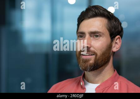 Close-up photo. Portrait of a successful and smiling young man with a beard and wearing a red shirt sitting in the office and confidently looking away. Thinks, dreams, poses. Stock Photo