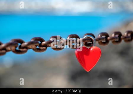 A single red, unmarked, heart shaped, love lock or love padlock attached to a thick chain by sweethearts to symbolise their affection or love Stock Photo