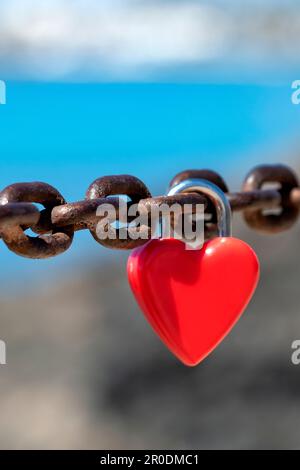 A single red, unmarked, heart shaped, love lock or love padlock attached to a thick chain by sweethearts to symbolise their affection or love Stock Photo