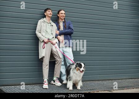 tattooed and cheerful gay man in casual outfit holding leash of Australian shepherd dog and standing next to positive boyfriend with pigtails hairstyl Stock Photo