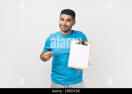 Portrait of smiling happy delighted unshaven man wearing blue T- shirt standing pointing at clipboard with empty paper, copy space. Indoor studio shot isolated on gray background. Stock Photo