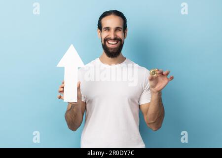 Portrait of smiling dark haired man with beard wearing white T-shirt showing growth increase of bitcoin, expressing positive emotions. Indoor studio shot isolated on blue background. Stock Photo