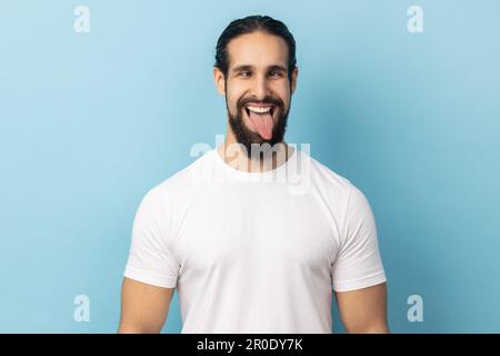 Portrait of comic positive man with beard wearing white T-shirt looking cross-eyed, having fun with silly face expression, showing tongue out. Indoor studio shot isolated on blue background. Stock Photo