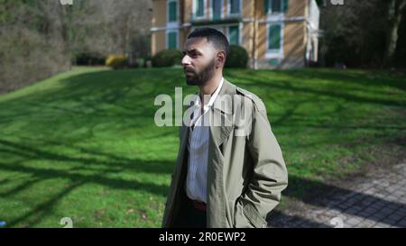 One confident young Middle Eastern man walking outside at park wearing fashionable jacket. An Arab male person with serious expression walks outdoors Stock Photo