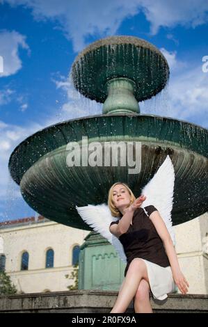 Mid adult woman wearing angel wings sitting near fountain at university, Munich, Bavaria, Germany Stock Photo