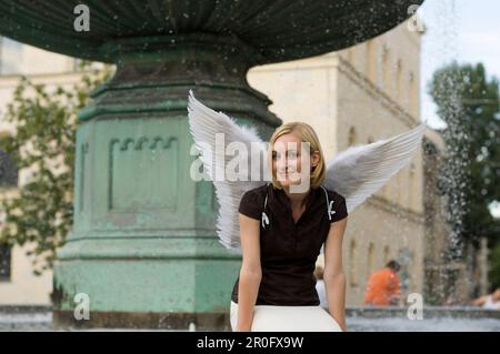 Mid adult woman wearing angel wings sitting near fountain at university, Munich, Bavaria, Germany Stock Photo
