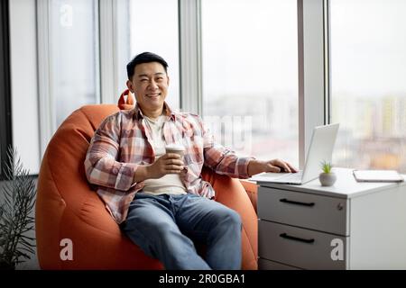 Portrait of casually dressed positive indian man, sitting on orange bean bag pouffe and smiling at camera in comfortable office. Smiling worker holding cup of aromatic coffee and typing on laptop. Stock Photo