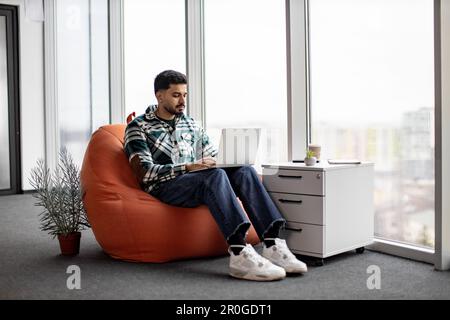 Casually dressed indian programmer working closely on writing and testing codes for new mobile applications in spacious glassy office. Focused man using personal laptop for doing particular job. Stock Photo