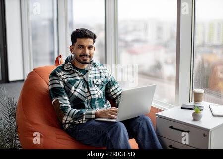 Portrait of nice-looking indian employee sitting on soft office chair with personal laptop on knees and smiling at camera. Imposing dark haired man taking short break after intensive working day. Stock Photo
