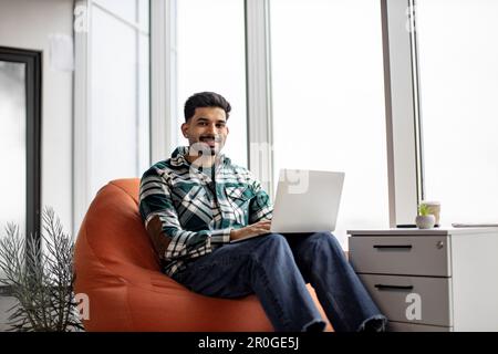 Portrait of nice-looking indian employee sitting on soft office chair with personal laptop on knees and smiling at camera. Imposing dark haired man taking short break after intensive working day. Stock Photo