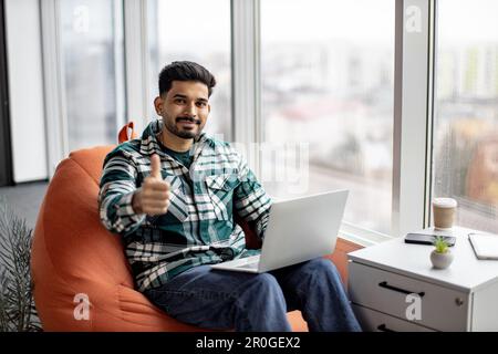 Smiling bearded indian man holding working laptop on knees and showing thumb up. Young attractive male sitting on bean bag chair near large panoramic window and posing on camera. Freelance concept. Stock Photo