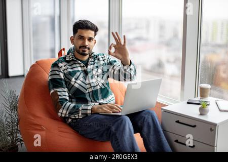 Smiling bearded indian man holding working laptop on knees and showing sign ok. Young attractive male sitting on bean bag chair near large panoramic window and posing on camera. Freelance concept. Stock Photo