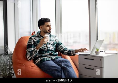 Good-looking indian man casually dressed sitting on soft bean bag chair with cup of tasty coffee and typing on wireless gadget. Young bearded male promoting new business plan in cozy workplace. Stock Photo